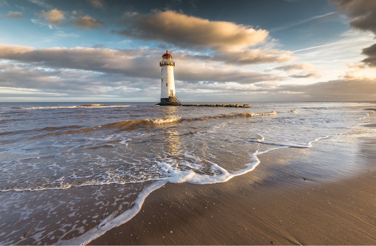 A lighthouse with waves coming up the sand on the beach and clouds in a blue sky.